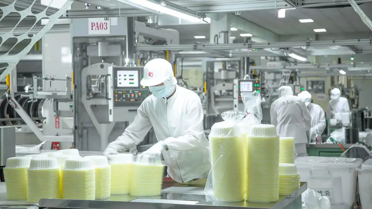 Workers in cleanroom gear packing stacked plastic food containers on a production line under food packaging certification hygiene controls